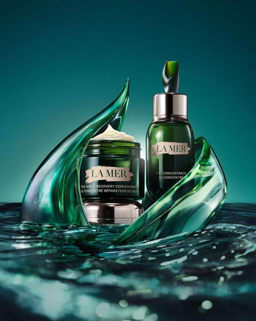 A visually striking display of La Mer skincare products, including a jar of The Night Recovery Concentrate and a bottle, surrounded by flowing green glass elements in a water-like setting.