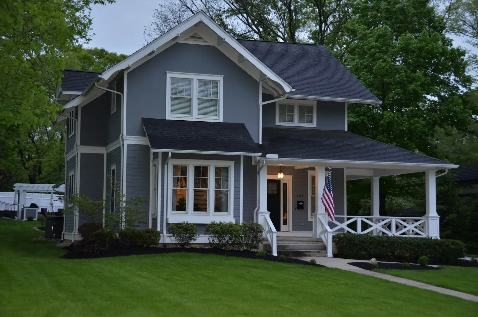 Two-story gray craftsman-style house with a covered front porch, white trim, manicured lawn, and surrounding trees.