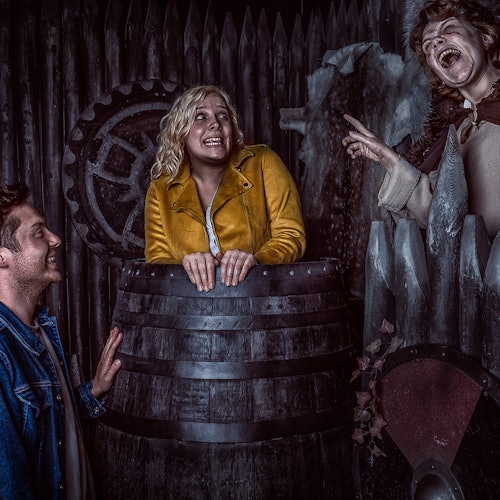 Three people in a dark, eerie setting; one in a barrel, another laughing on a ledge, and the third smiling in a denim jacket.