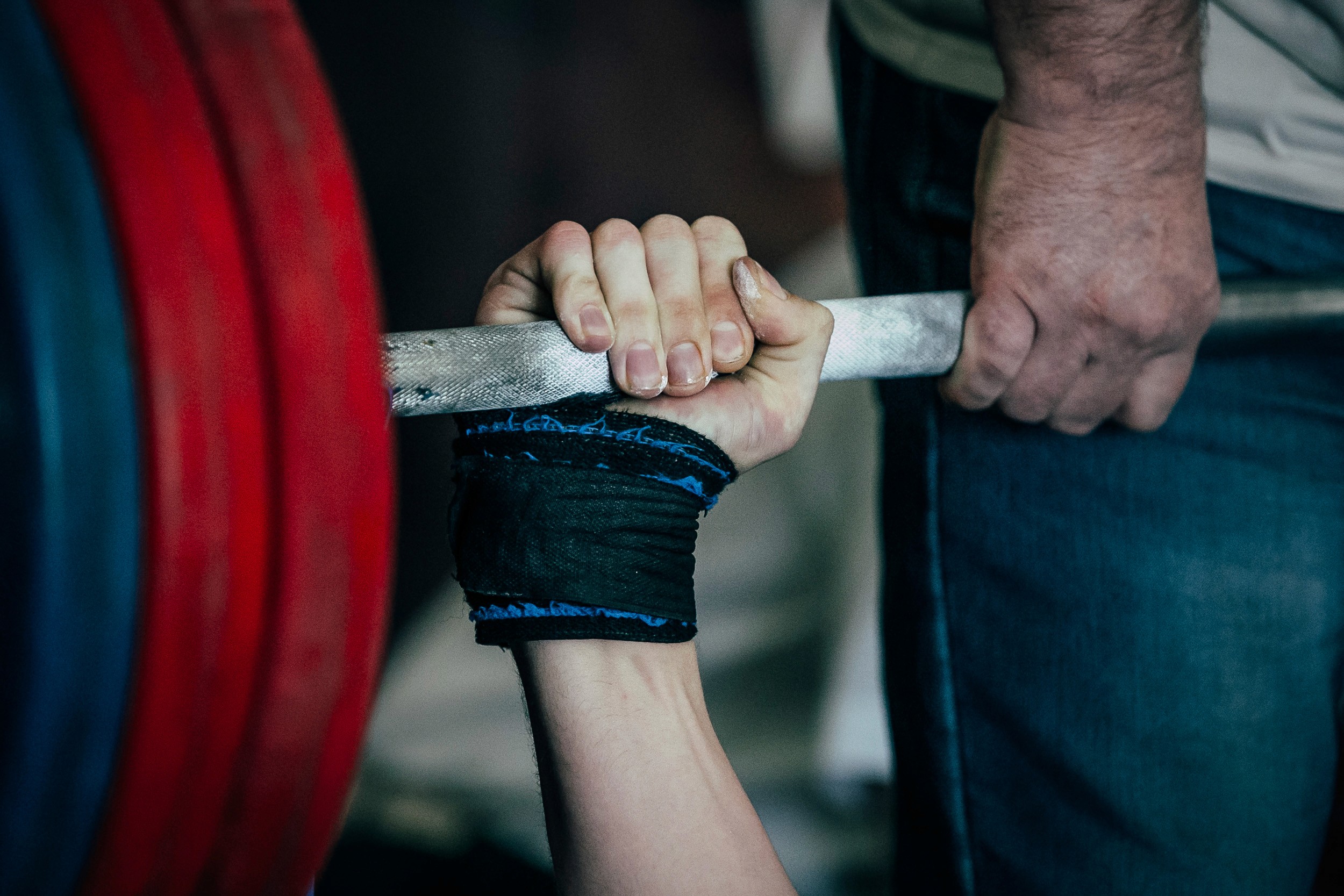 A close-up shot of a person's hands during a bench press exercise. One hand is gripping the barbell, showing the use of wrist wraps for support, while the other hand, likely belonging to a spotter, is positioned nearby to assist. The barbell is loaded with large weight plates in red and blue, emphasizing the heavy weight being lifted. The image captures the tension and teamwork involved in weightlifting, with clear details of the person's muscular hand and the rough texture of the barbell.