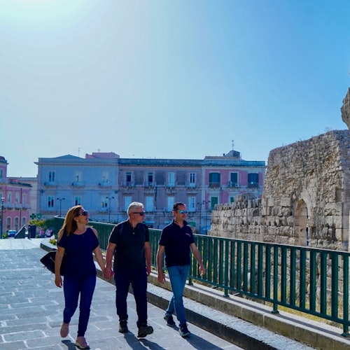 Three people walk along a stone-paved path beside ancient ruins and pastel-colored buildings under a clear blue sky.