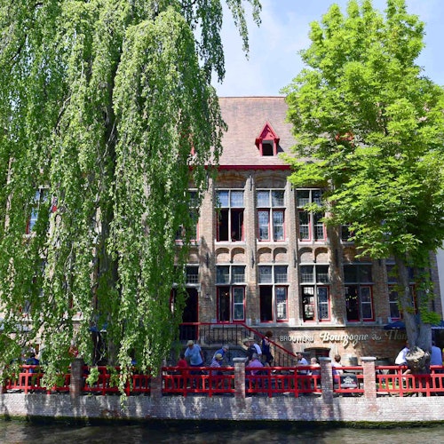 Folk sitter på en utendørs kafe med rødt gjerde, ved en kanal, foran en historisk bygning med høye vinduer og frodige trær.