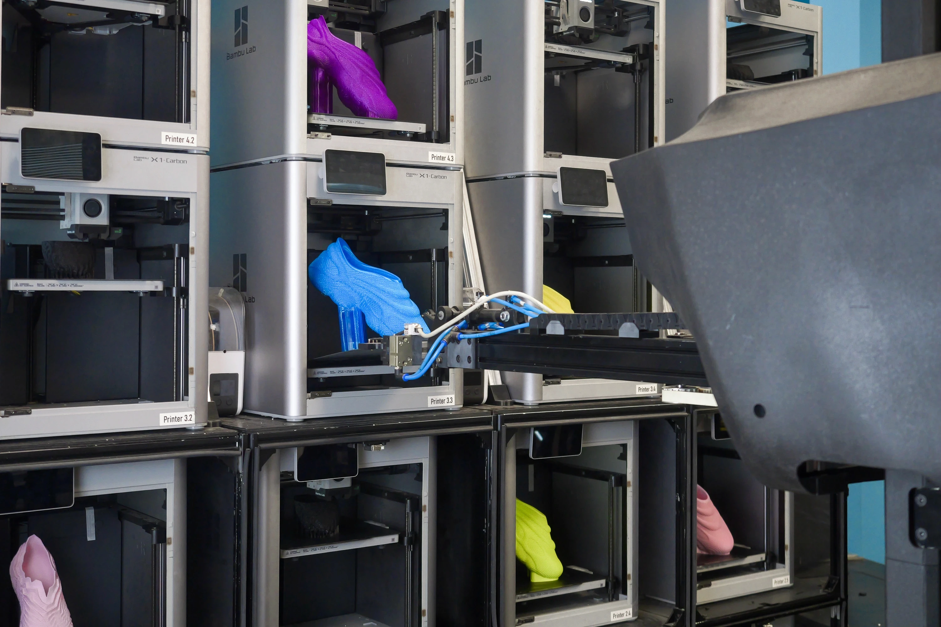 A footwear print farm with stacked 3d printers and a robot arm, performing an automated build plate removal from a printer.