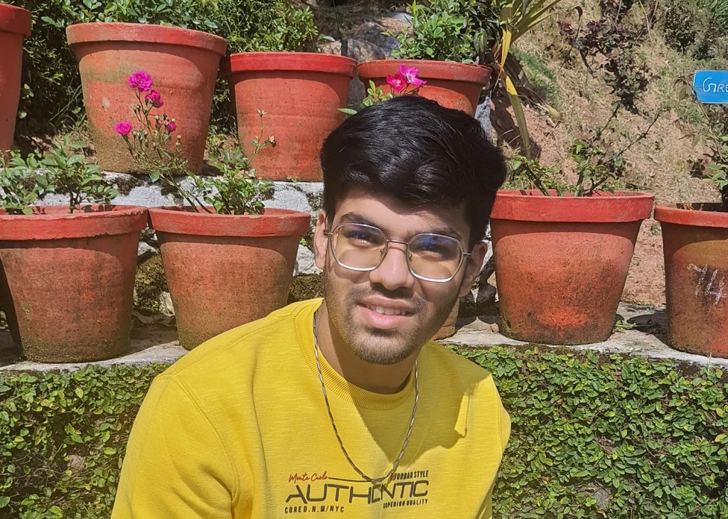 A person wearing headphones sits at a desk with a large curved monitor displaying graphic design software, surrounded by a laptop, smartphone, and potted plant, creating a modern workspace setup.