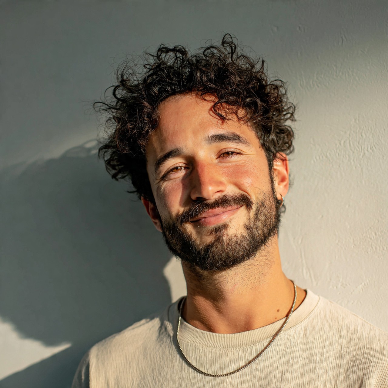 Young man with curly hair and beard smiling in sunlight.