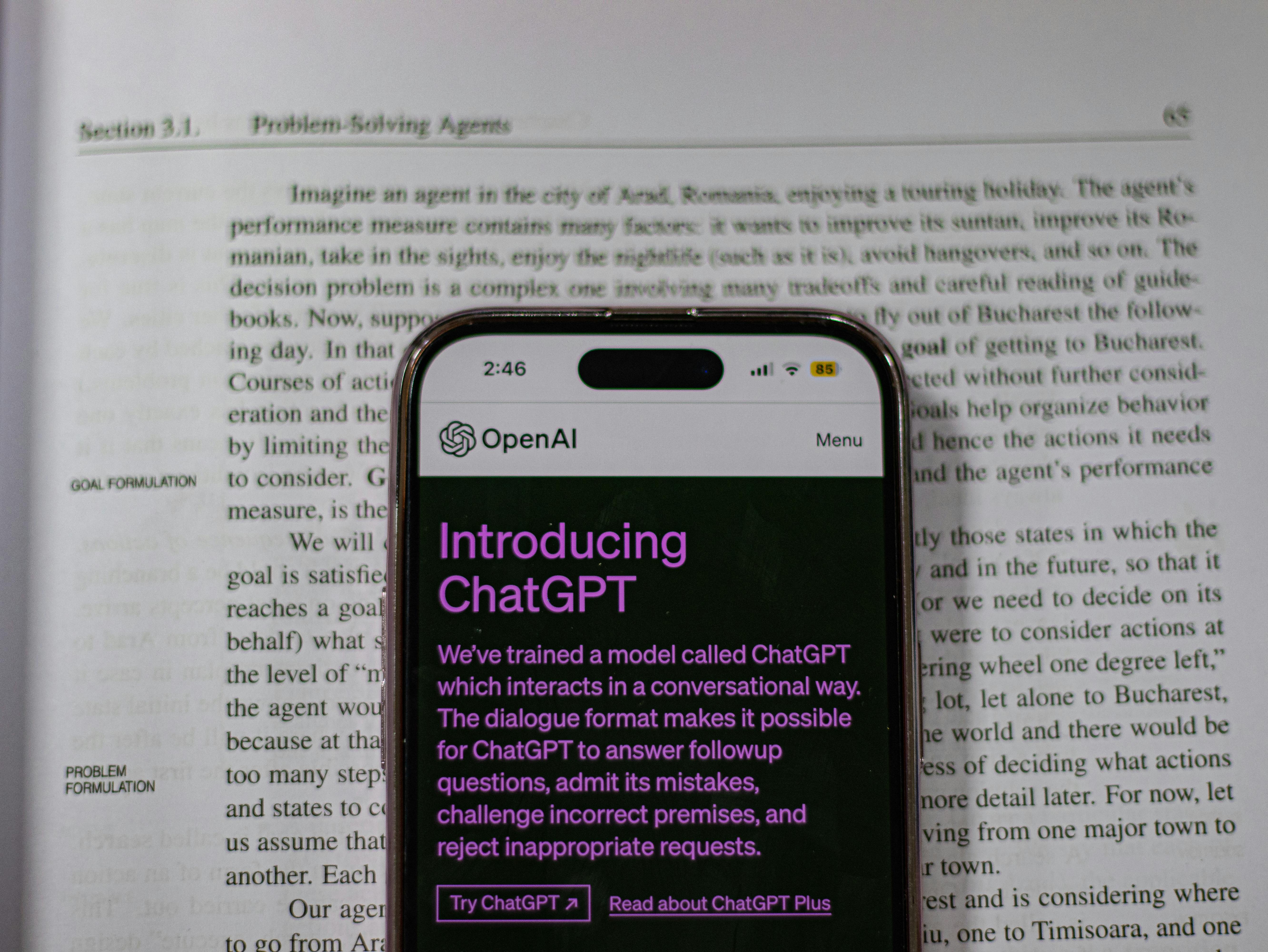 A smartphone displaying OpenAI's ChatGPT introduction page is placed over an open book. The phone shows text about ChatGPT in purple on a dark background.