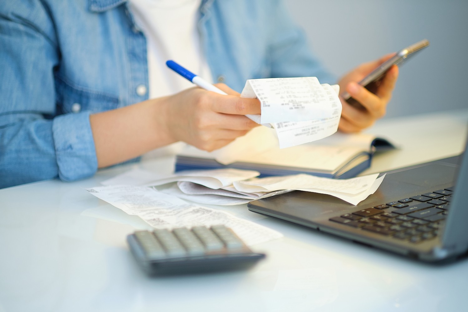 Person reviewing documents and writing notes with a laptop and calculator on the desk, representing care staffing agency compliance review