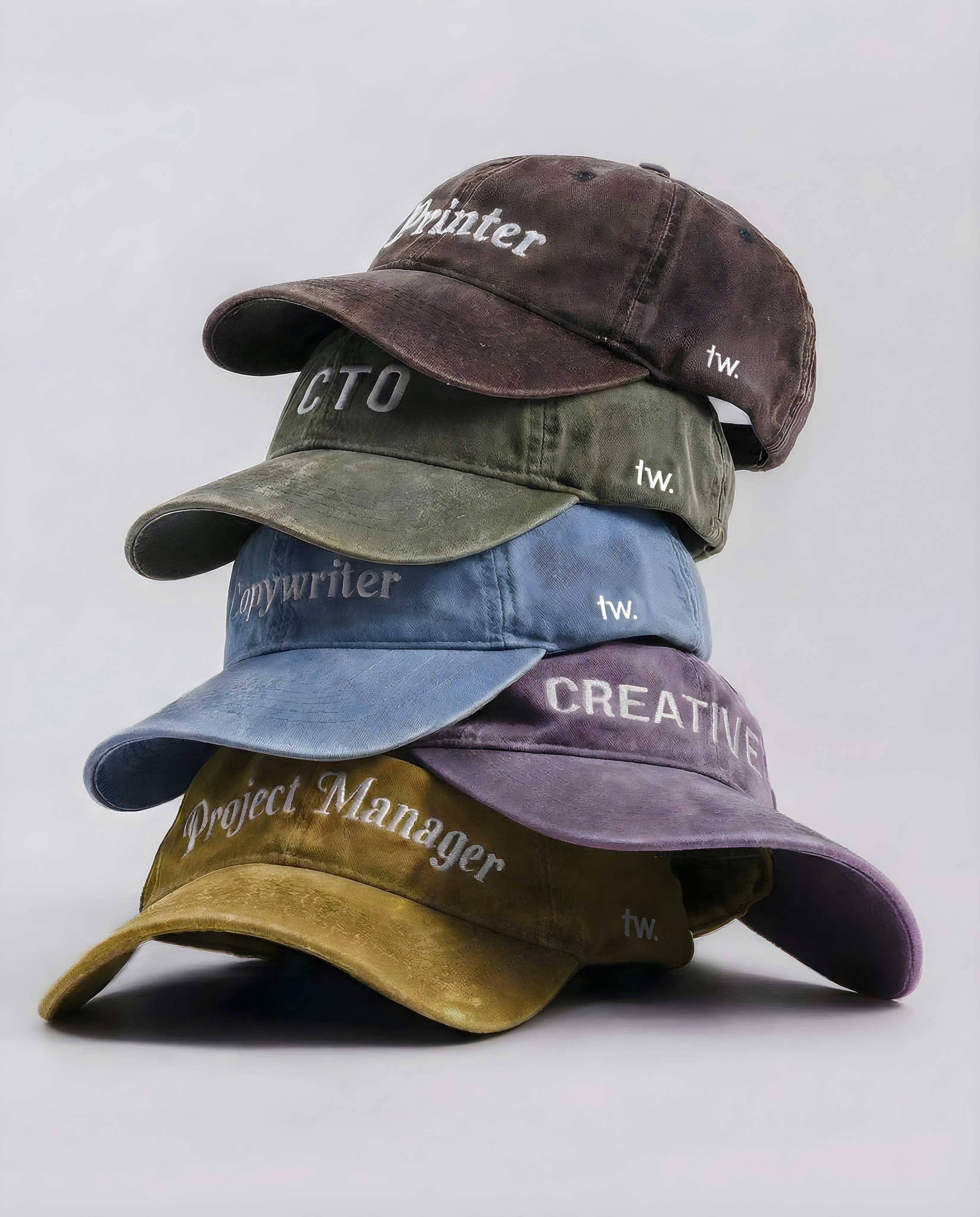 A stack of five worn cotton baseball caps in different colours, each embroidered with an agency role on the front and a small “tw.” mark on the side, photographed on a pale grey background.