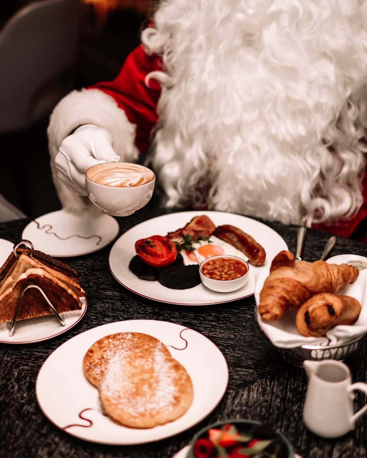 Santa enjoying a festive Christmas breakfast at Dhoom, featuring pancakes, croissants, toast and a cappuccino, capturing the warm and joyful holiday dining atmosphere.