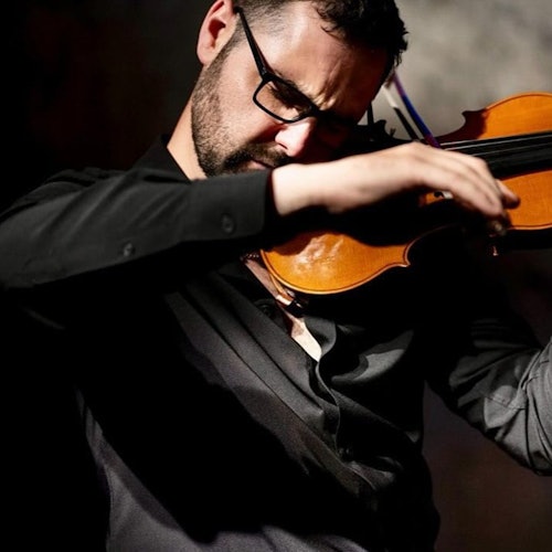 A person wearing glasses and a black shirt playing a violin with focus, set against a dark, blurred background.