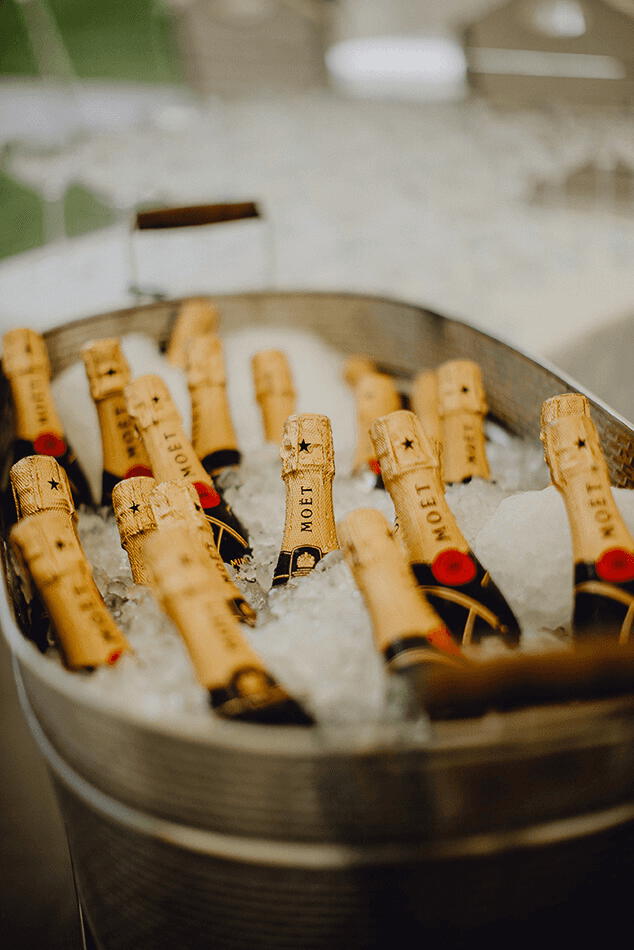 Outdoor restaurant terrace overlooking a tranquil lake at sunset. White tables and chairs are set with glassware. Sheer curtains add elegance. Calm ambiance.