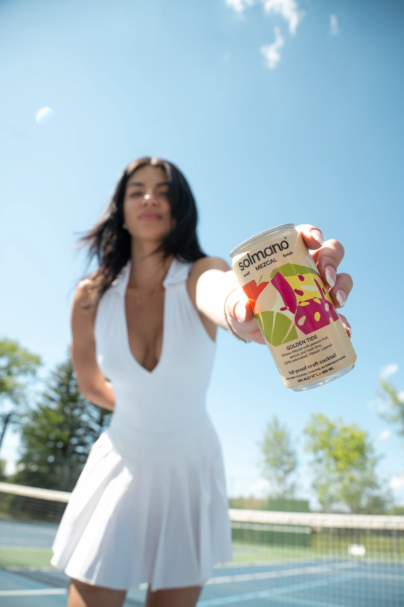 Person holding a Solmano Golden Tide cocktail can overhead against a blue sky in a tennis court.