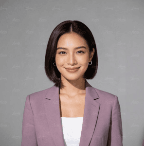 Smiling woman with a dark bob haircut wearing a purple blazer and hoop earrings against a gray background.