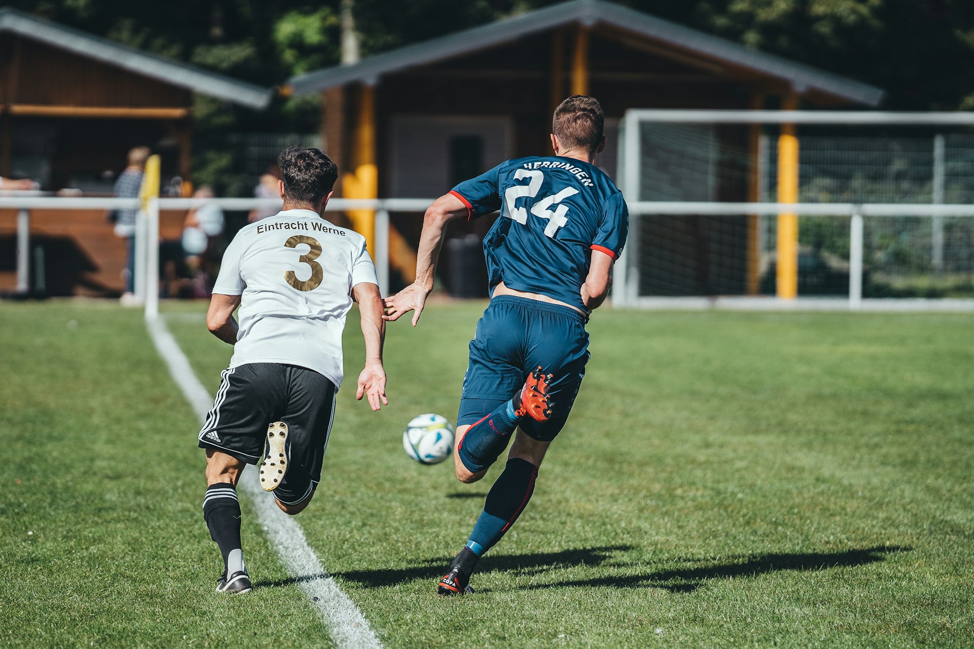 Two soccer players, one in a blue jersey and one in white, sprint side-by-side down a grass field chasing the ball.