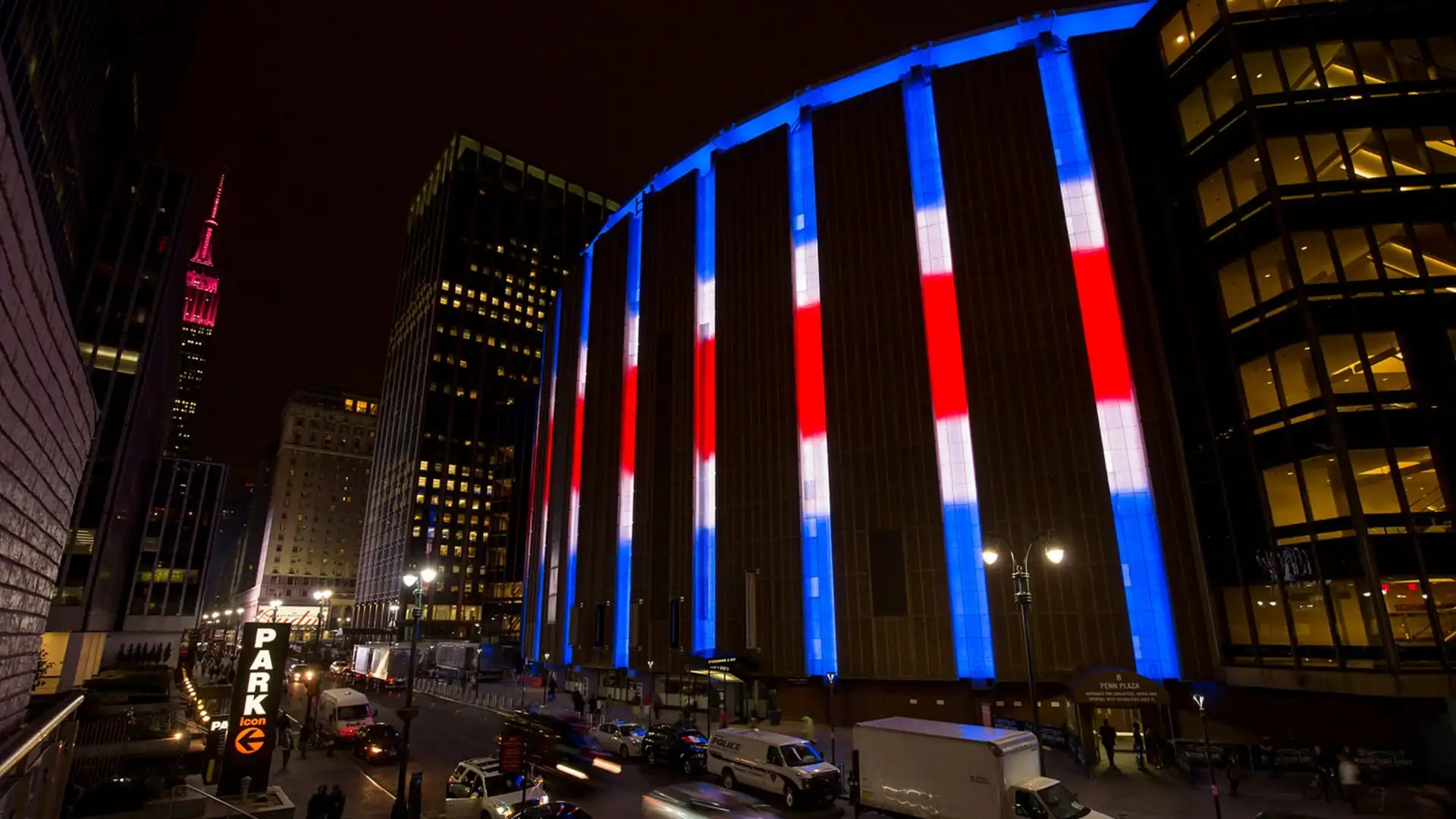 Exterior of Madison Square Garden at night lit up in red, white, and blue lights with the Empire State Building glowing in the background.