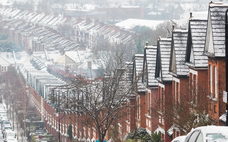 Rows of brick terraced houses stretch along a hillside, with rooftops and streets lightly covered in snow. Bare trees and parked cars line the neighbourhood, creating a wintry urban scene.