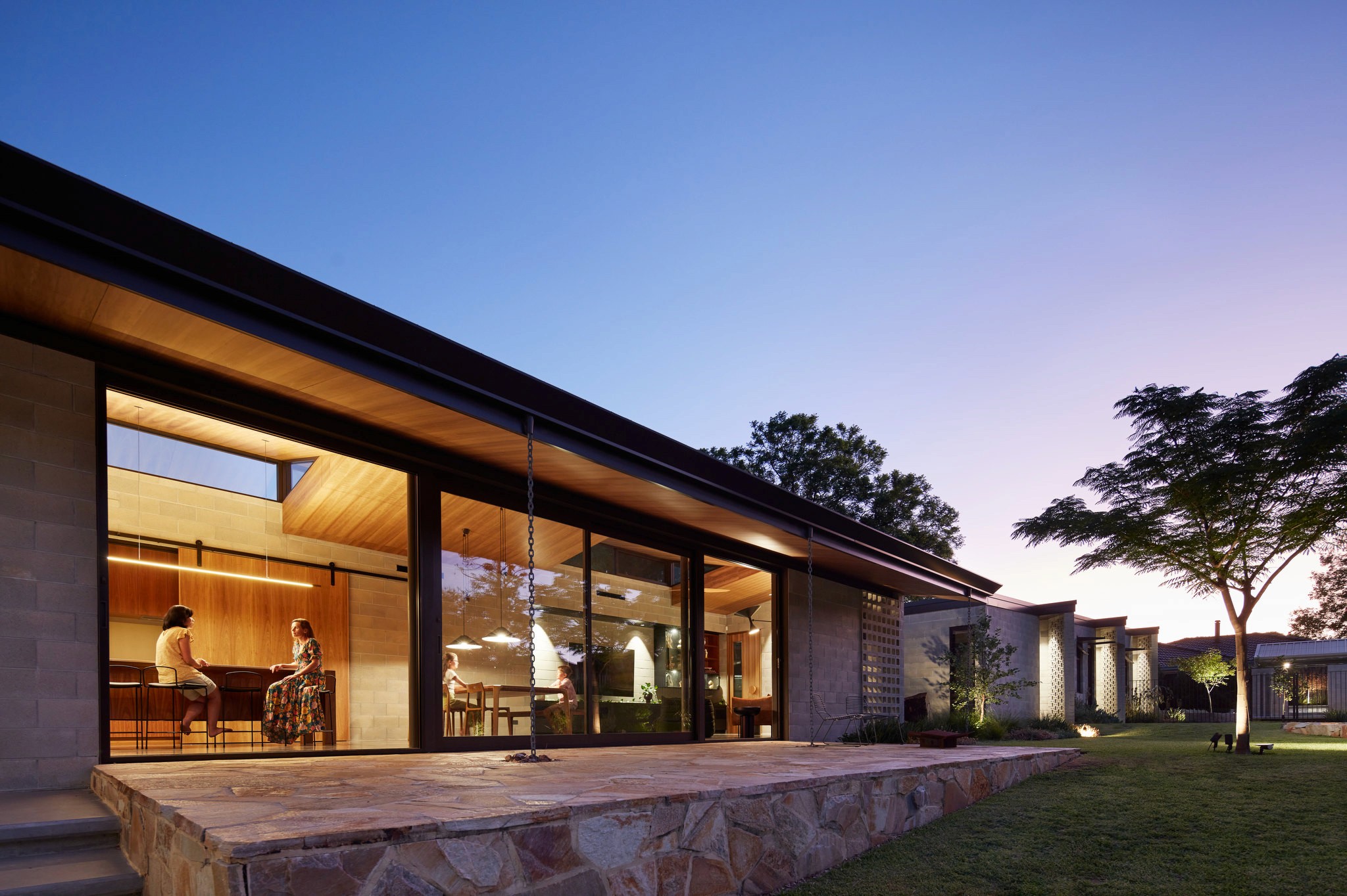 Modern glass and wood cabin with a cozy outdoor deck featuring wicker furniture, surrounded by lush green trees and a serene forest setting.
