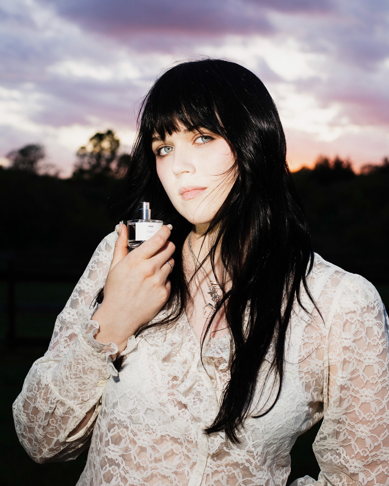 A woman with long black hair and bangs holds a SINGE Poet perfume bottle near her face, wearing a white lace blouse against an outdoor sunset sky.