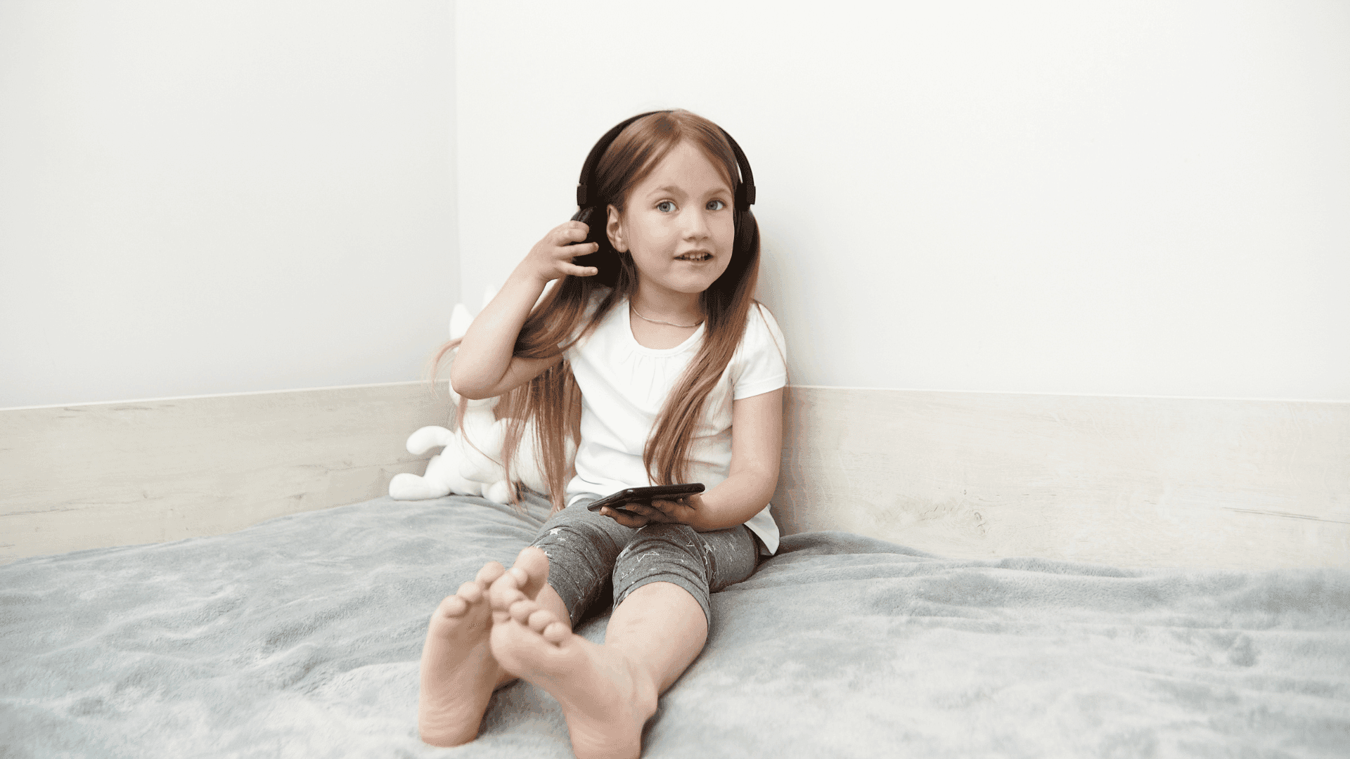 A young girl sitting on a grey blanket, wearing headphones and holding a phone, smiling while listening to HushAway®’s Sound Sanctuary.