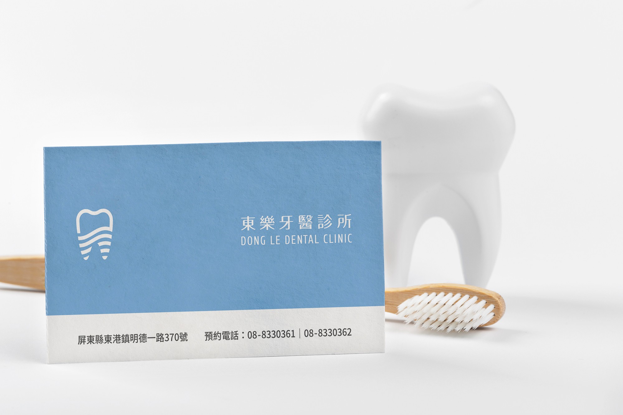 這張圖像展示了東樂牙醫診所的名片,名片為藍白色設計,上方有牙齒圖案和「東樂牙醫診所」及「DONG LE DENTAL CLINIC」的字樣,下方有地址和預約電話。