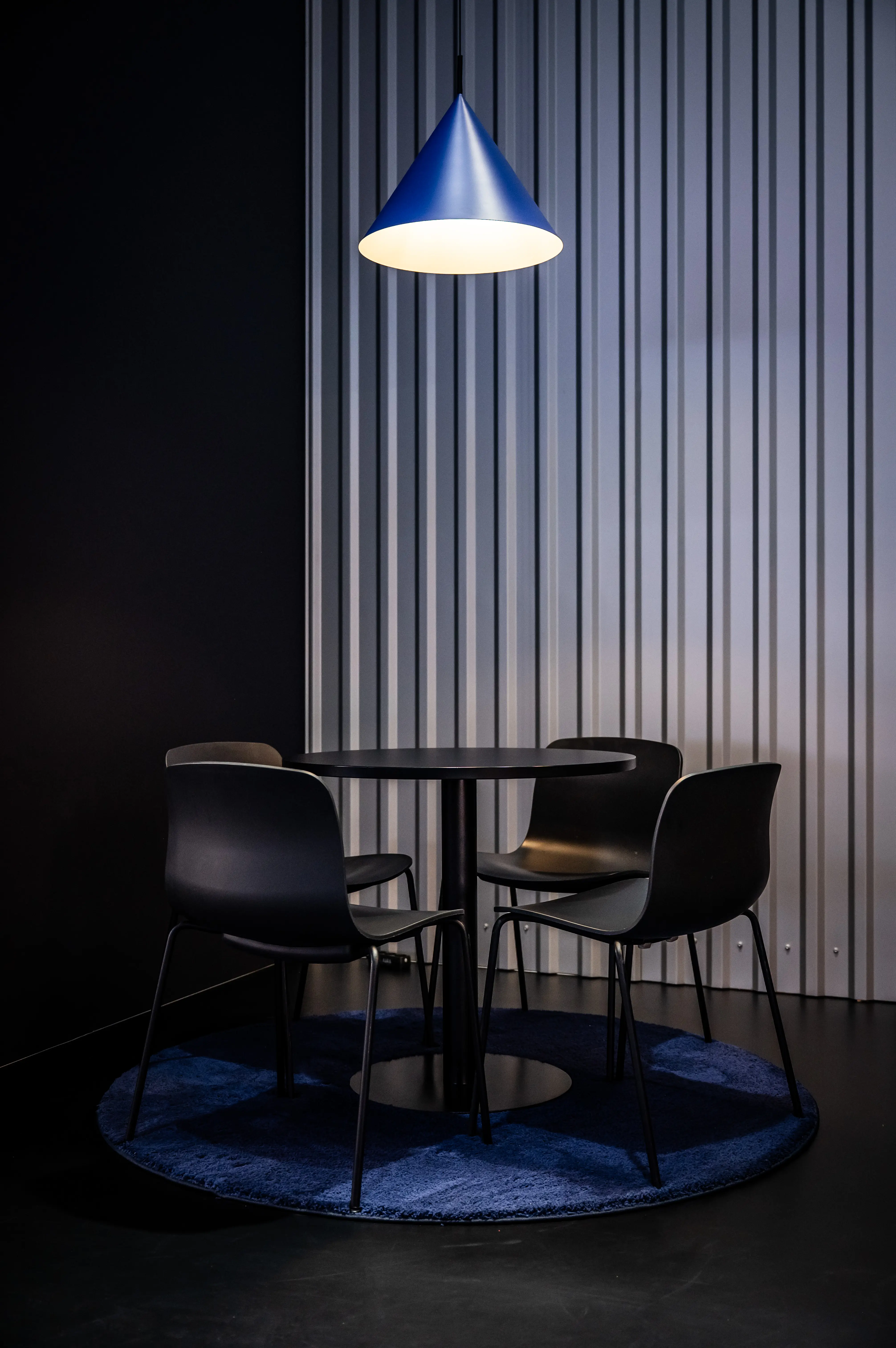Black round table with four chairs on a blue rug under a blue cone pendant light at the Revolut booth.