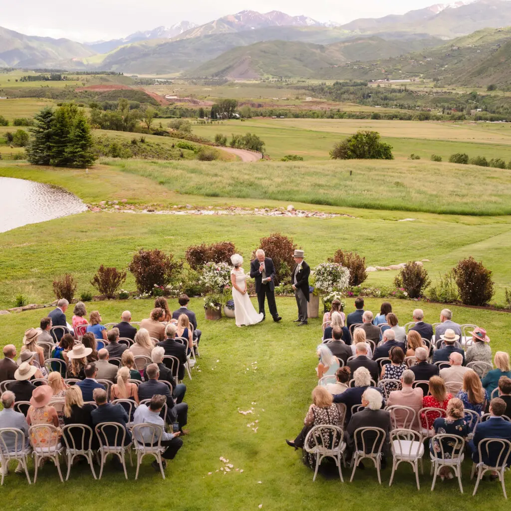 A high-angle wide shot of an outdoor wedding ceremony, showing rows of guests seated on a lush green lawn overlooking a mountain valley at a Chaparral Ranch Wedding.