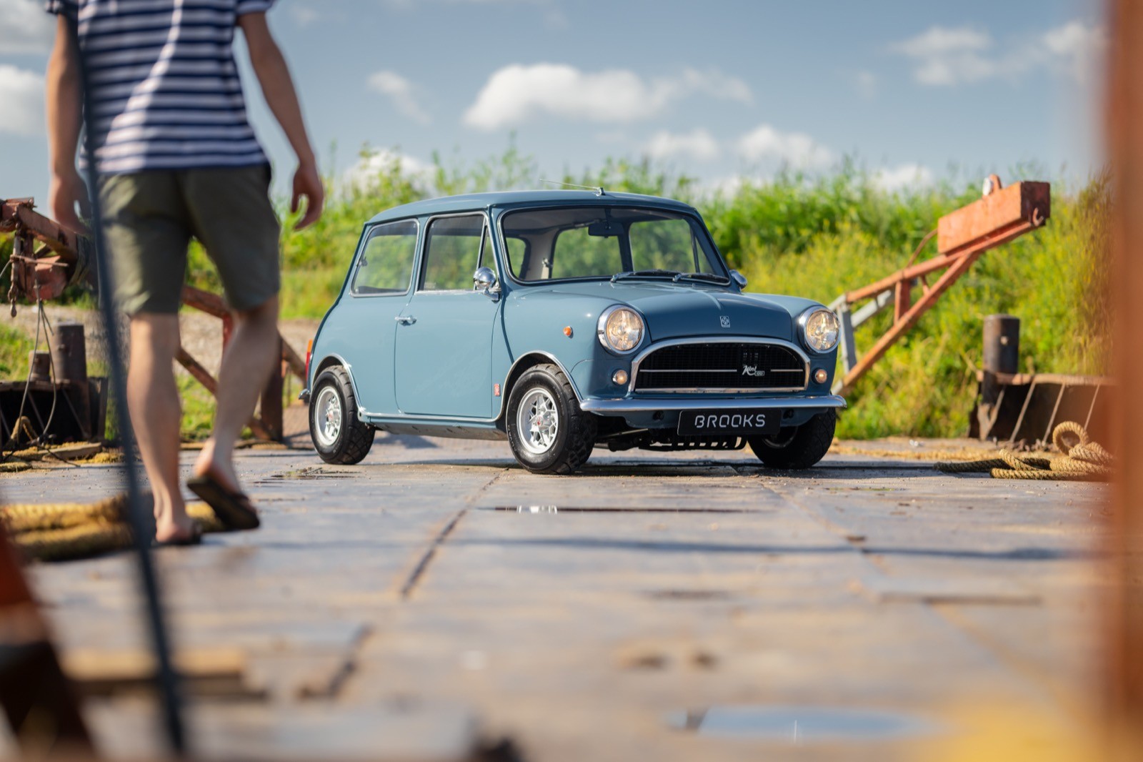 A vintage blue car parked on a wooden platform, with a person walking in the background and greenery around.