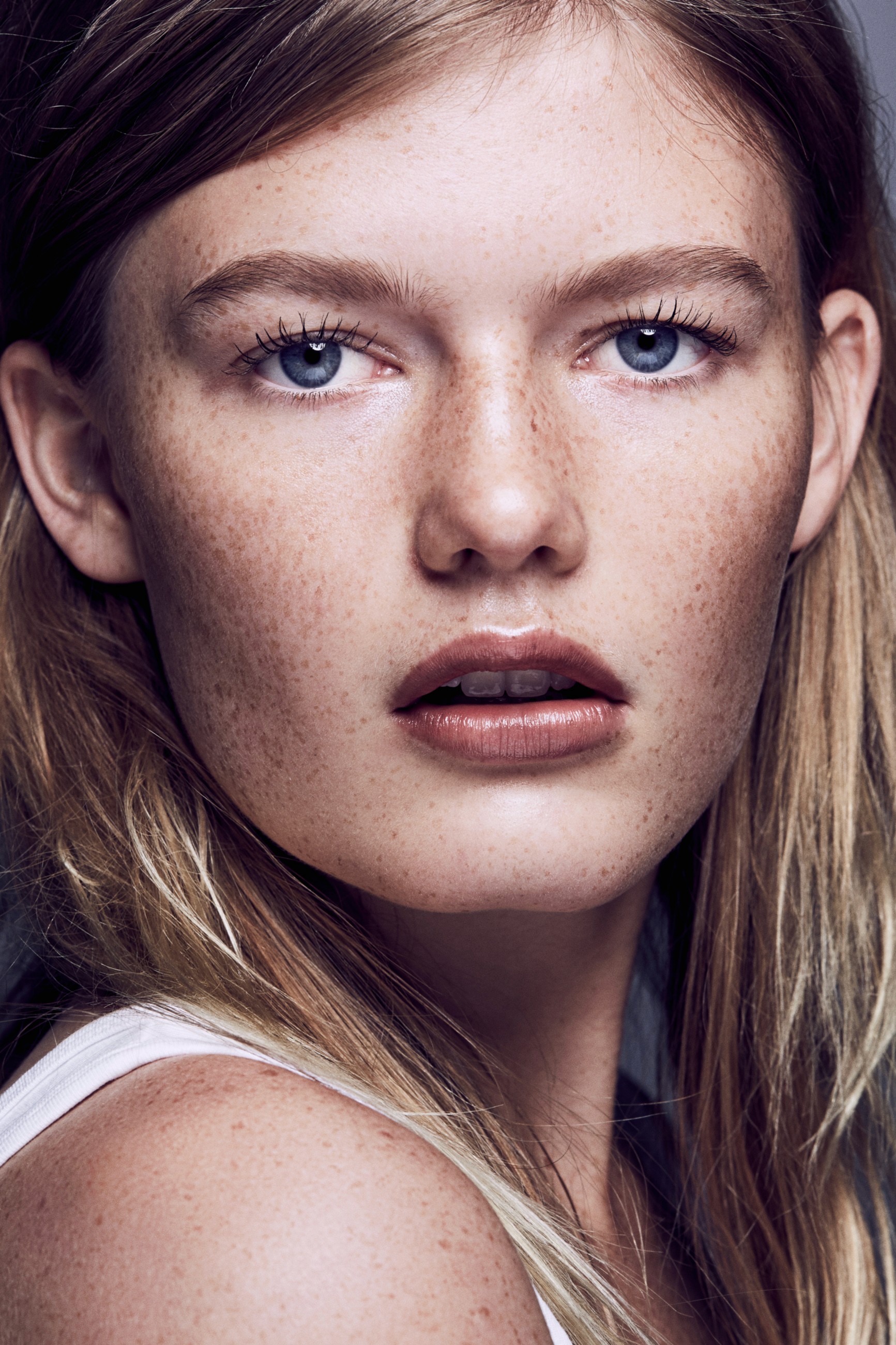 A close-up portrait of a model with freckly skin in beauty lighting looking at camera across her turned shoulder with blue eyes and a white singlet.