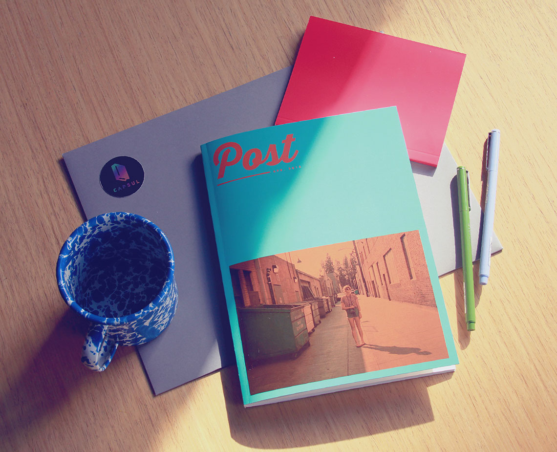 The image shows a wooden desk with a teal magazine titled "Post" lying flat on top of a gray envelope. A blue and white patterned mug rests beside the envelope. Two pens, one light green and one white, are placed near the magazine. A red note is positioned partially underneath the magazine. The scene is lit by natural light coming from the upper left.