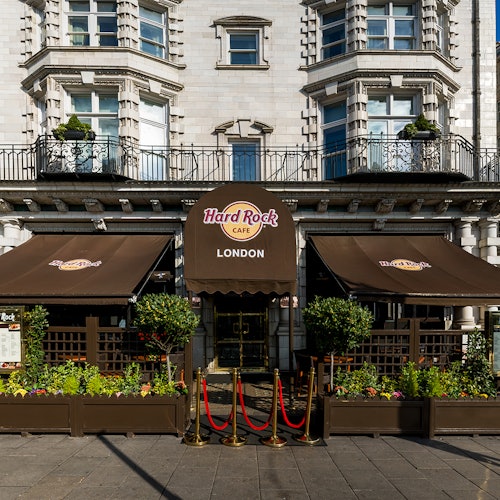Exterior of Hard Rock Cafe London with brown awnings, potted plants, and red ropes leading to the entrance.