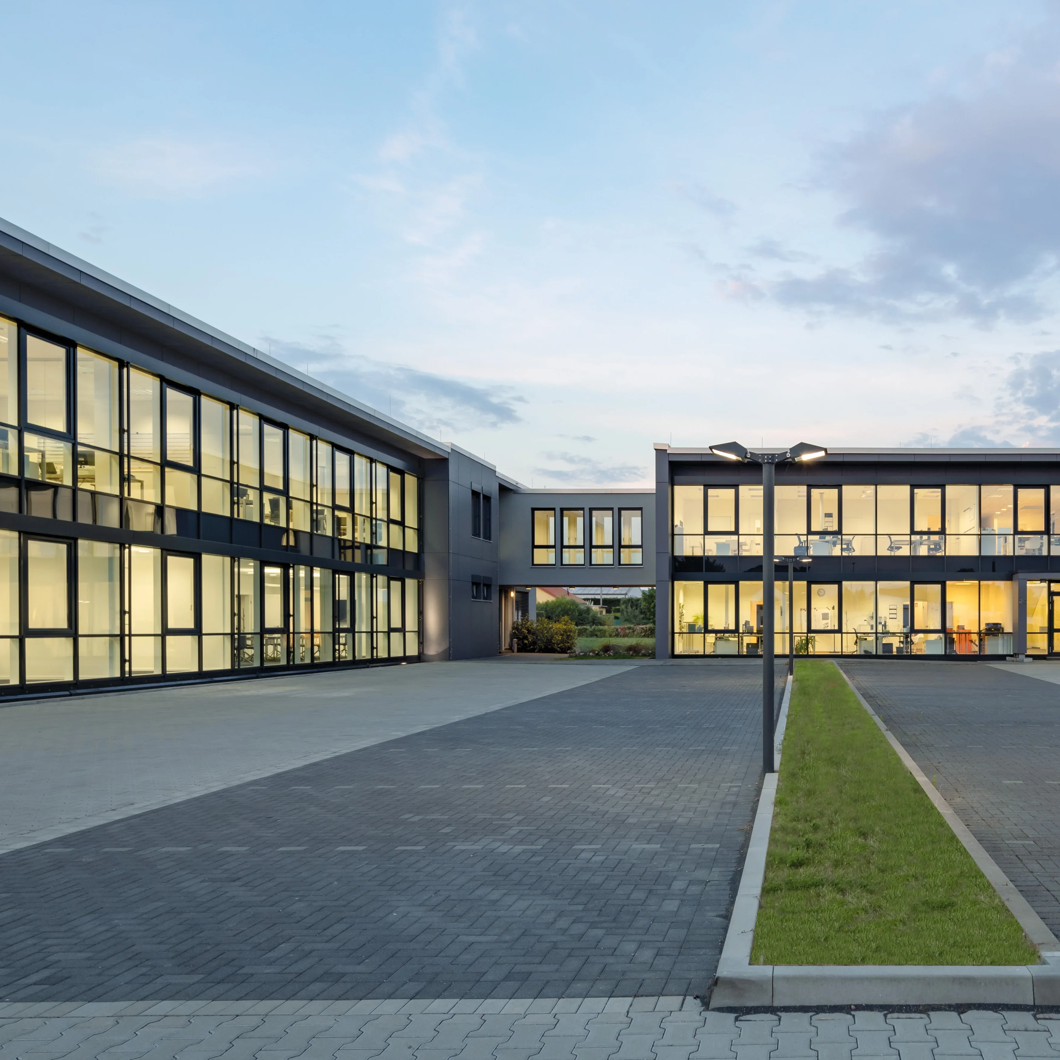 Moderner Gebäudekomplex mit großen Glasfronten, beleuchteter Innenraum und gepflasterter Hoffläche bei Abenddämmerung.