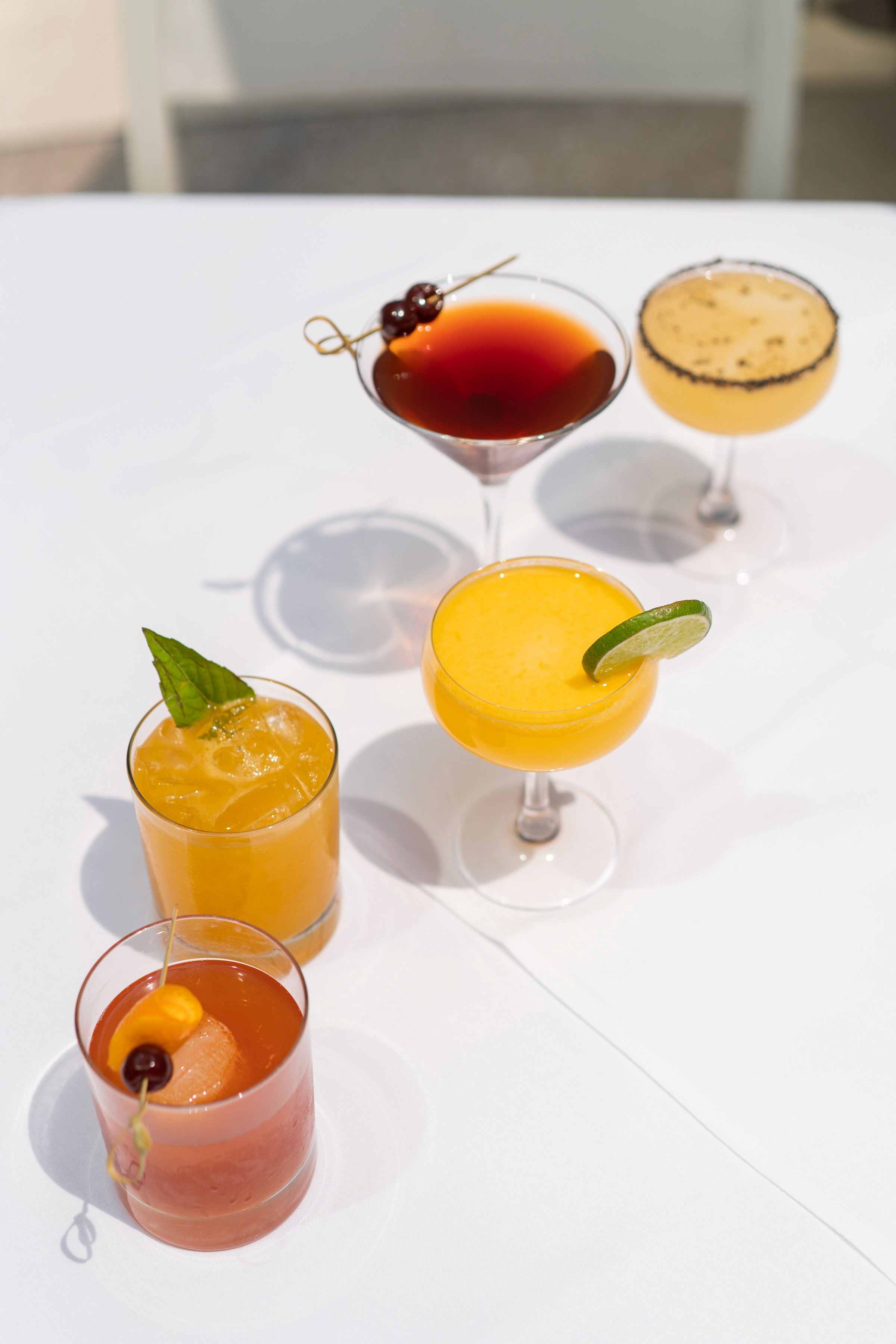 A line of colorful cocktails sits on a white table, featuring various garnishes and glassware.