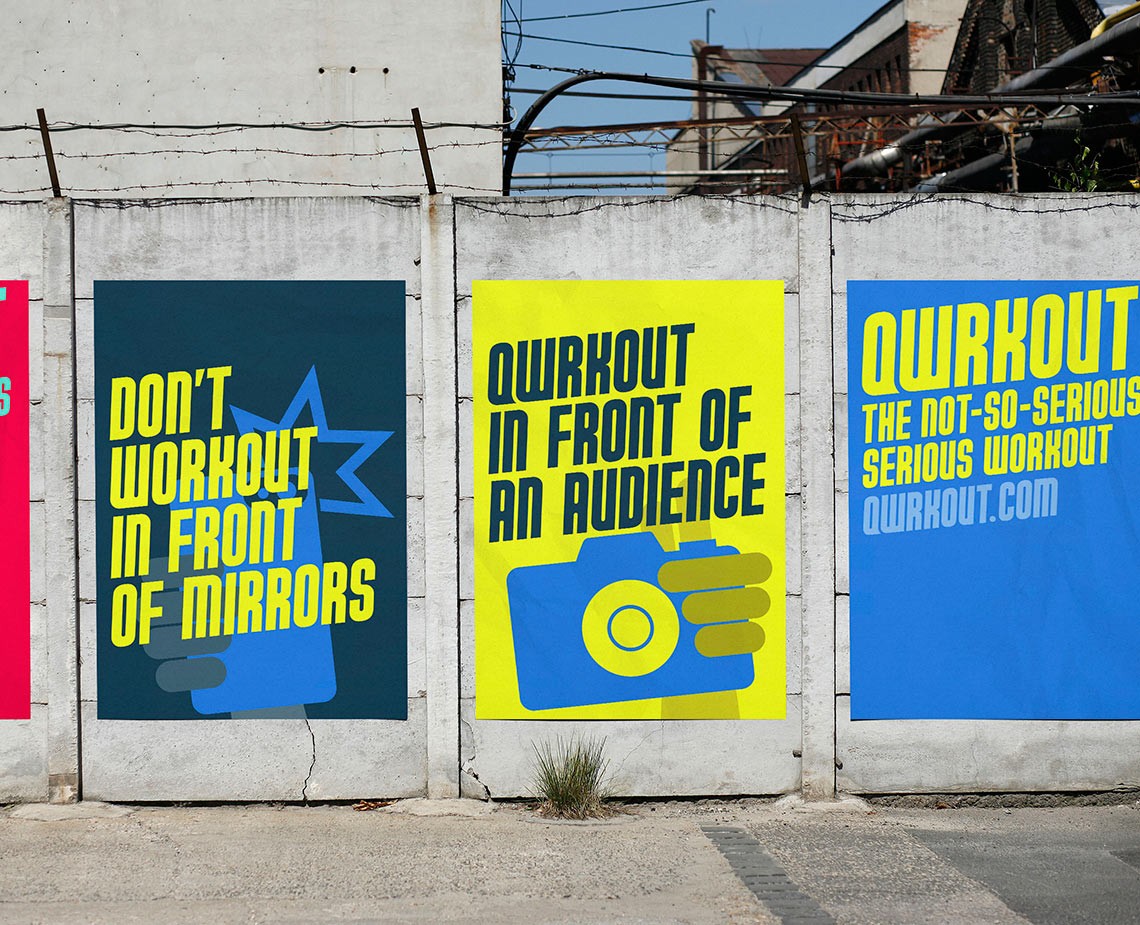 Three vertical banners on a concrete wall display motivational messages. The leftmost banner reads “DON’T WORKOUT IN FRONT OF MIRRORS.” The center banner reads “QURKOUT IN FRONT OF AN AUDIENCE.” The rightmost banner reads “QURKOUT THE NOT-SO-SERIOUS SERIOUS WORKOUT” with the website address “QURKOUT.COM” displayed below.