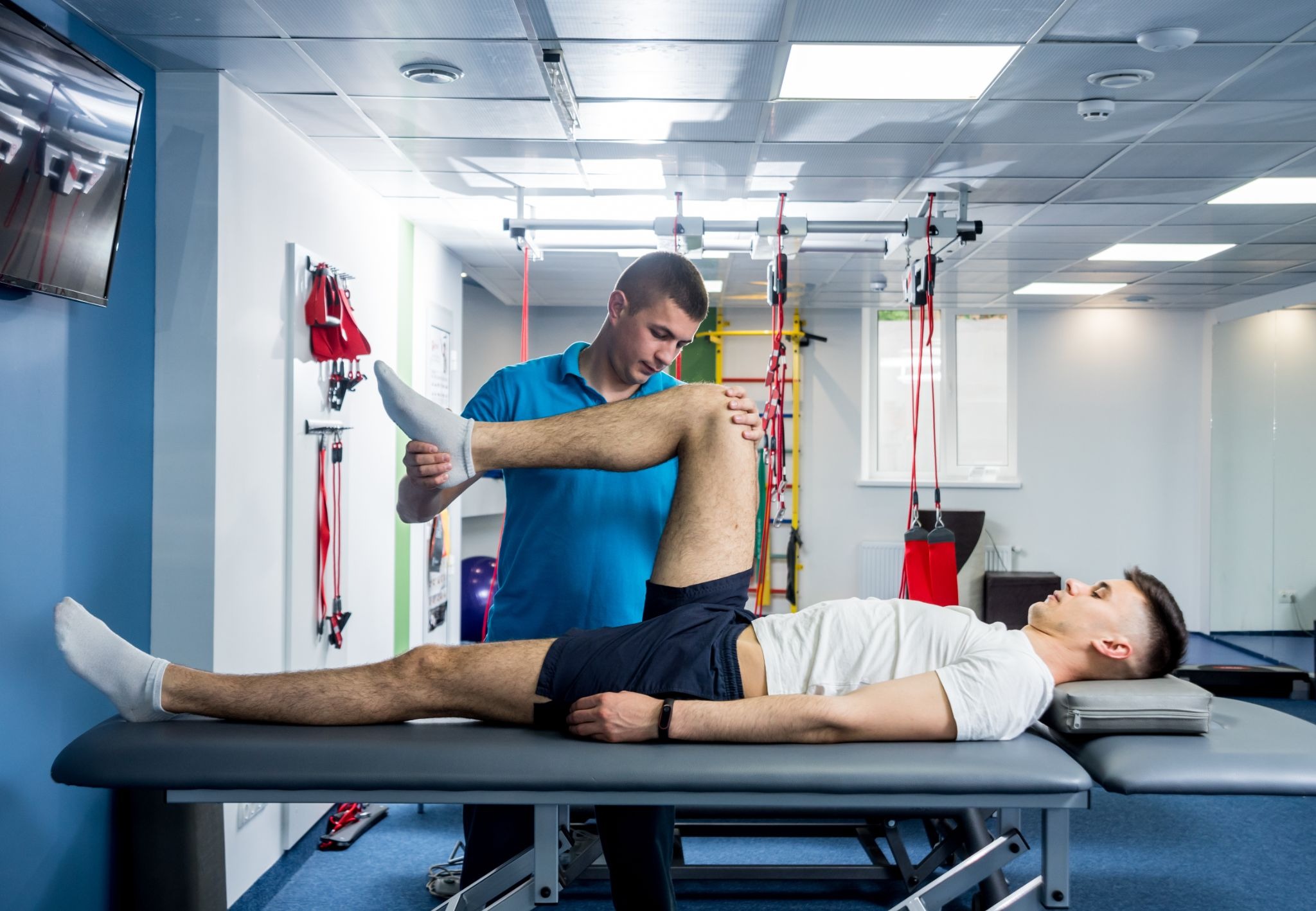 A physical therapist in a blue shirt guides a patient's leg through a range of motion stretch on a treatment table.