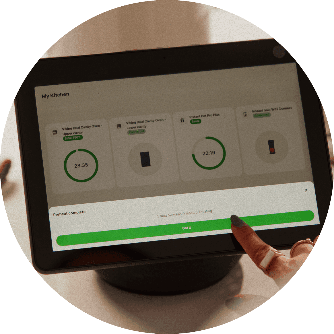 Tablet displays "My Kitchen" dashboard with multiple connected appliances showing timers like "28:35" and "22:19" while a user taps a green status bar reading "Preheat complete"