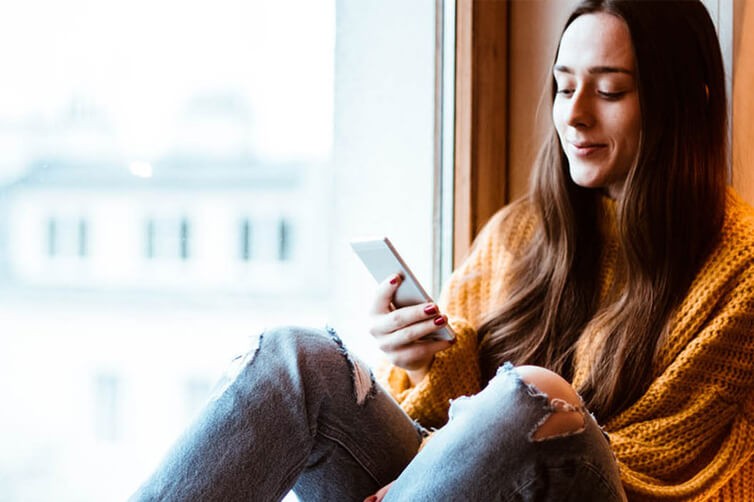Young woman in sweater using smartphone by window