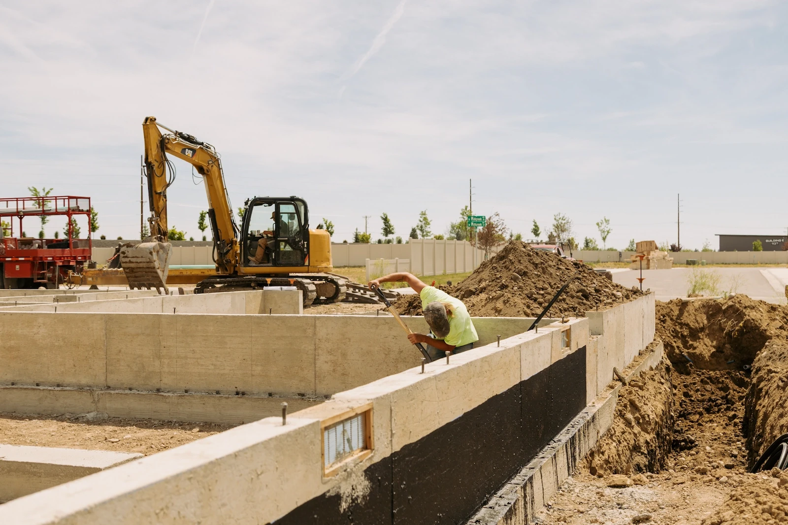 A worker leans over a poured concrete foundation wall while a yellow Caterpillar excavator operates in the background at a residential construction site.
