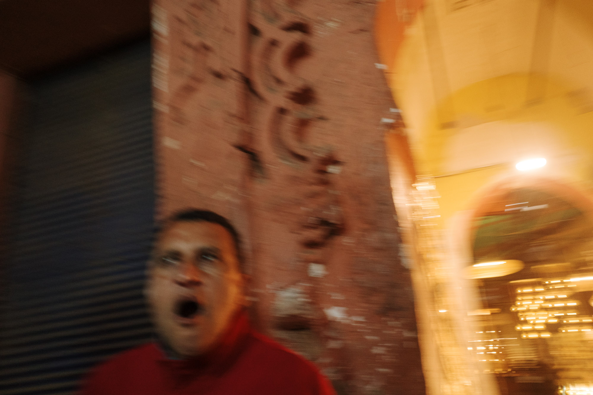 An individual expressing surprise stands in front of a blurred, brightly lit entrance, set against a textured, reddish-brown wall.
