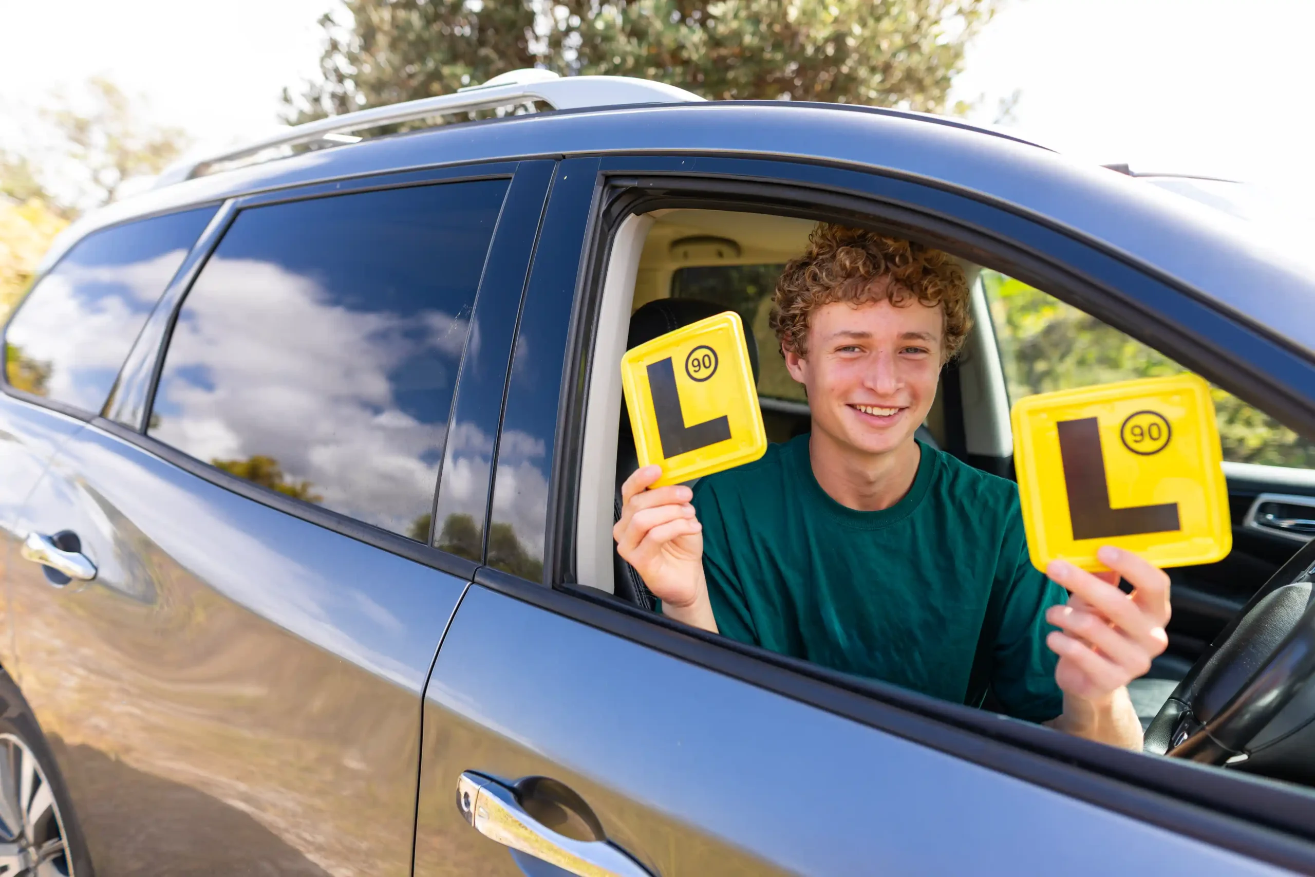 Young adult in the drivers seat of a vehicle, smiling, holding L plates.