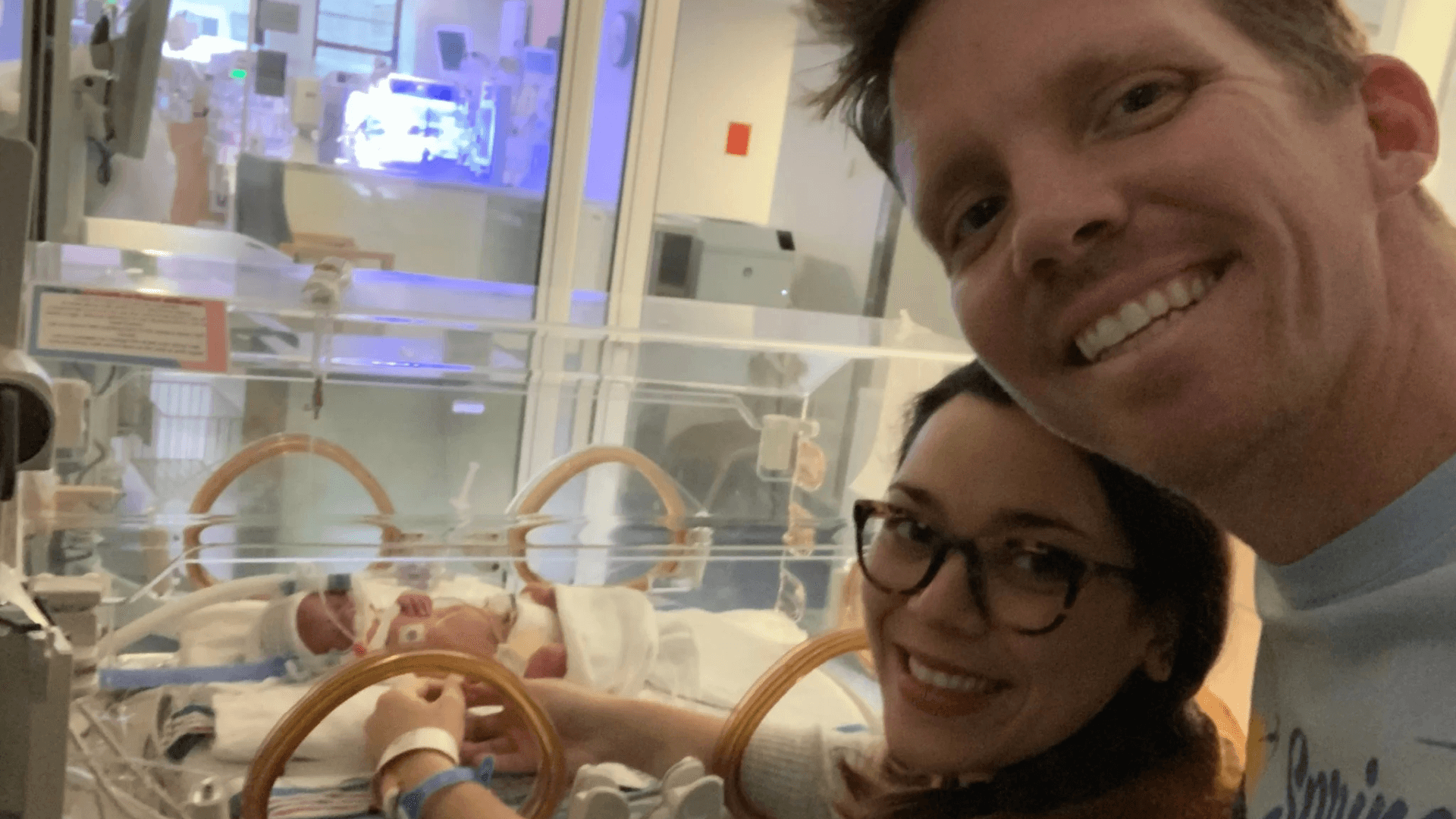 Smiling parents taking a selfie beside their newborn baby resting in a hospital incubator in a neonatal intensive care unit, with medical equipment visible in the background.
