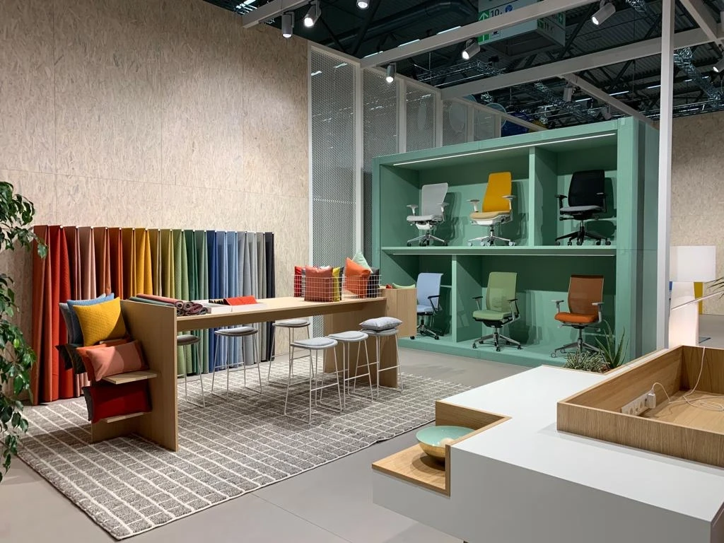 Inside the HAWORTH ORGATEC stand with a mint-green shelf of office chairs in many colors, a wooden table with stools and a colorful felt panel divider.