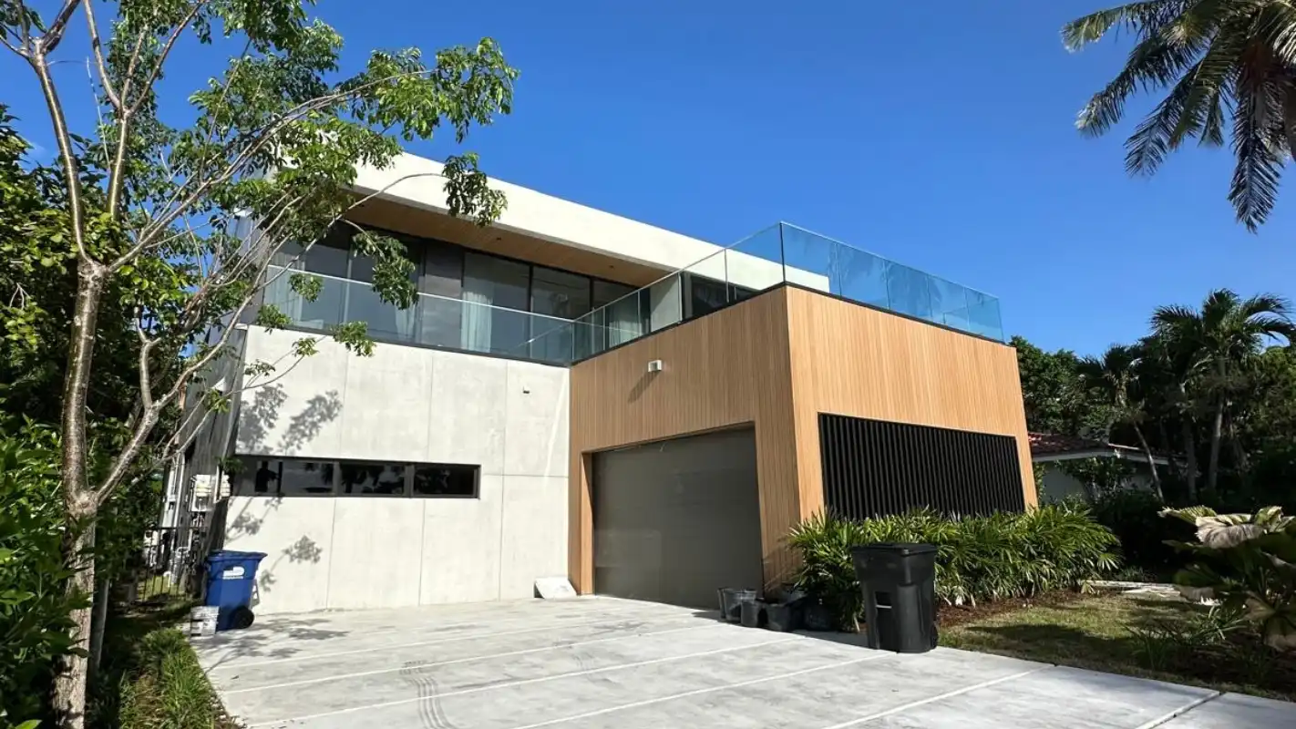 Modern Miami home exterior with wood and concrete facade, glass rooftop railing, and a wide driveway lined with palms