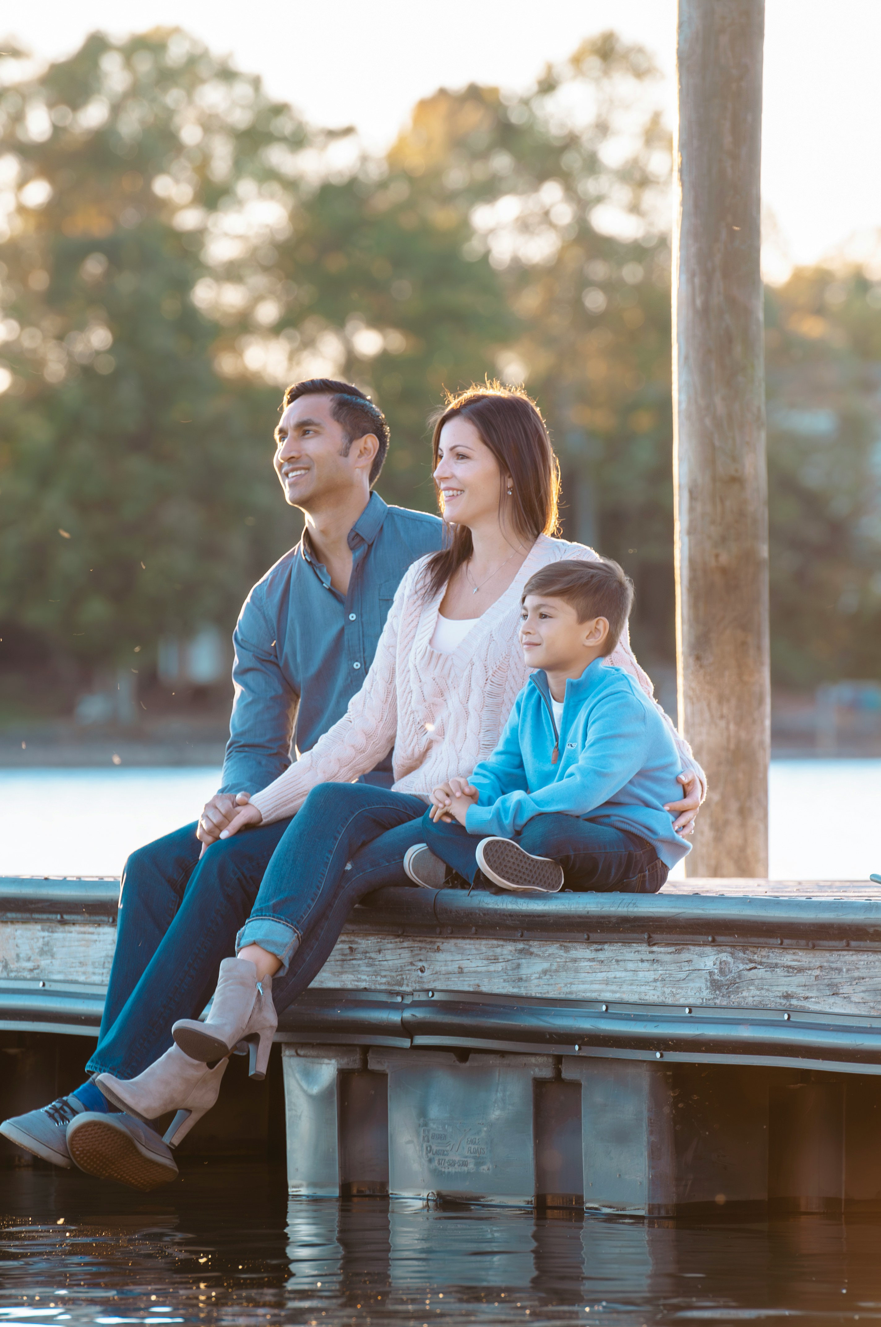 A family sits together on a wooden dock by a serene lake, enjoying a sunny day, with lush green trees in the background, creating a peaceful and happy scene.