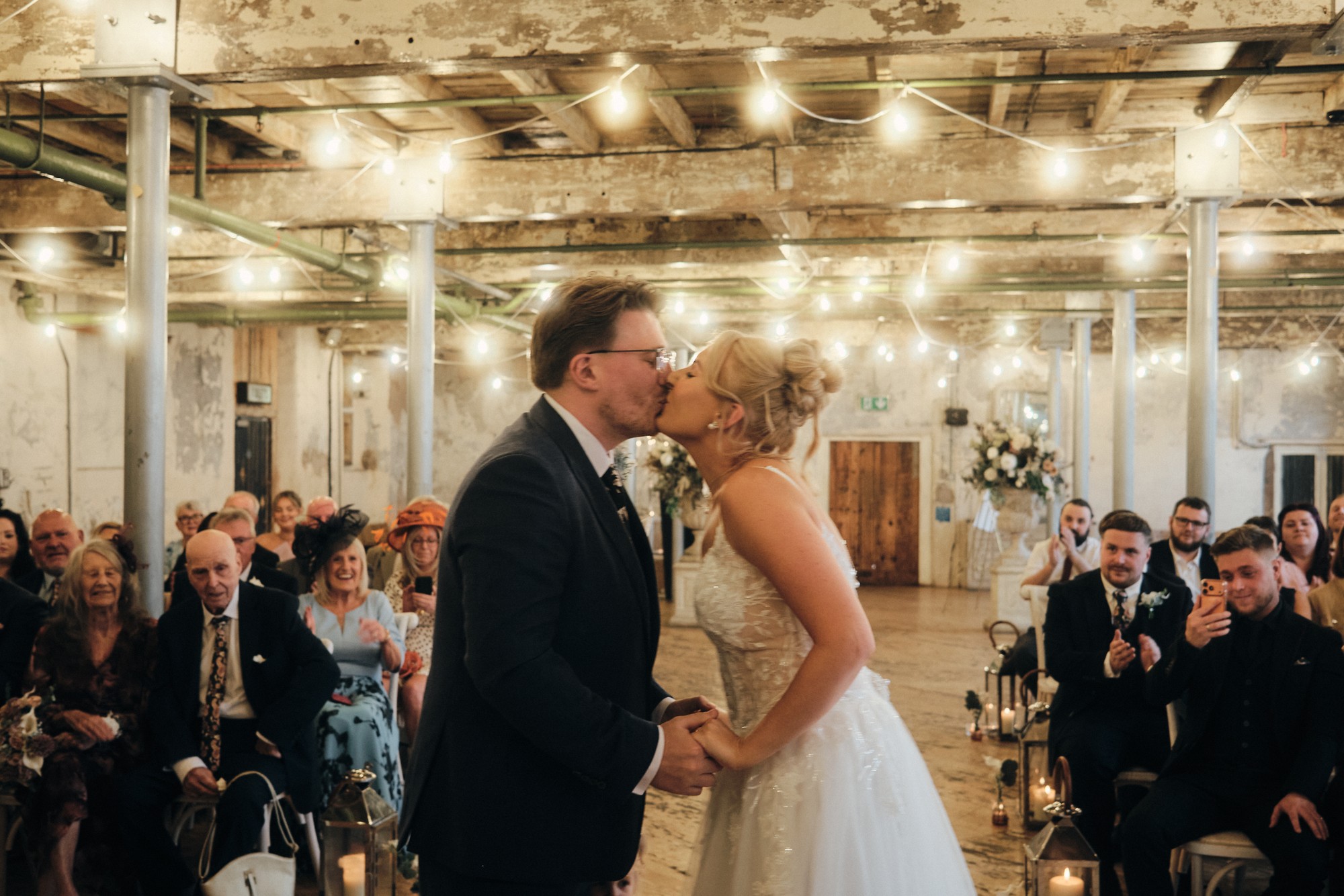 A bride and groom kissing during their ceremony at Holmes Mill, with guests applauding beneath strings of festoon lights