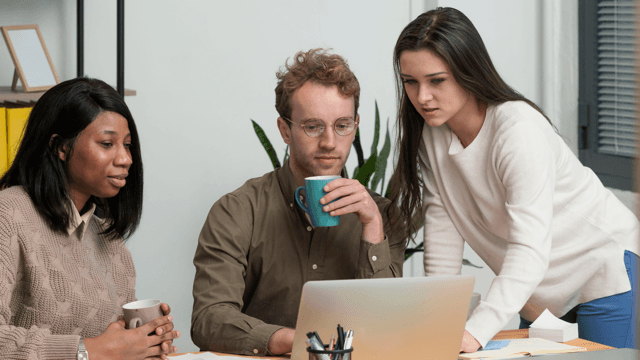 Photo of a man and a lady seated and looking into a laptop with another lady looking over their shoulder