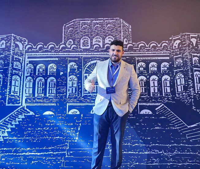 Man sitting in a dimly lit room, wearing a beige shirt and looking contemplative.