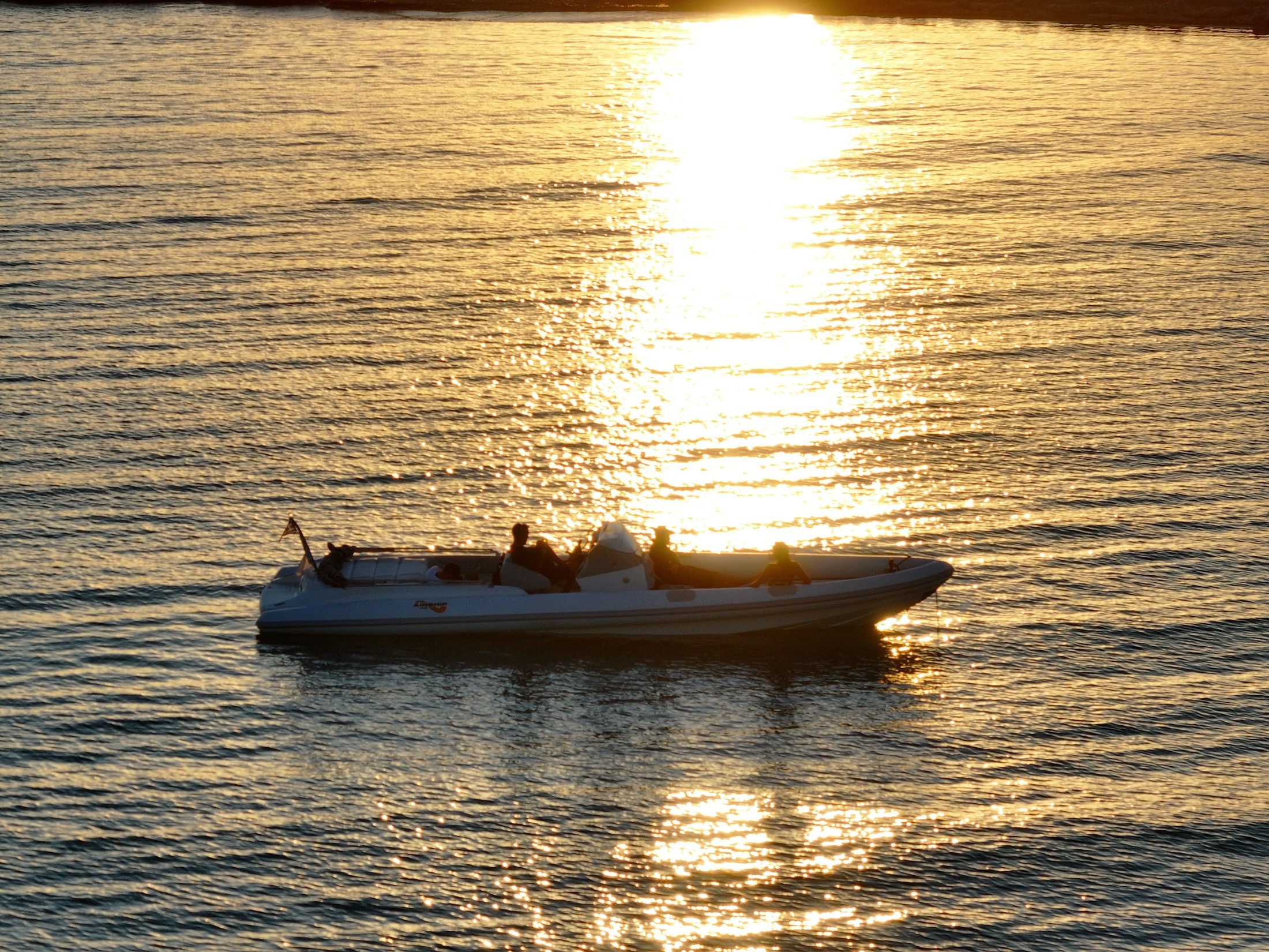 White speedboat with passengers cruising on golden waters during sunset, creating a shimmering reflection path across the calm sea.