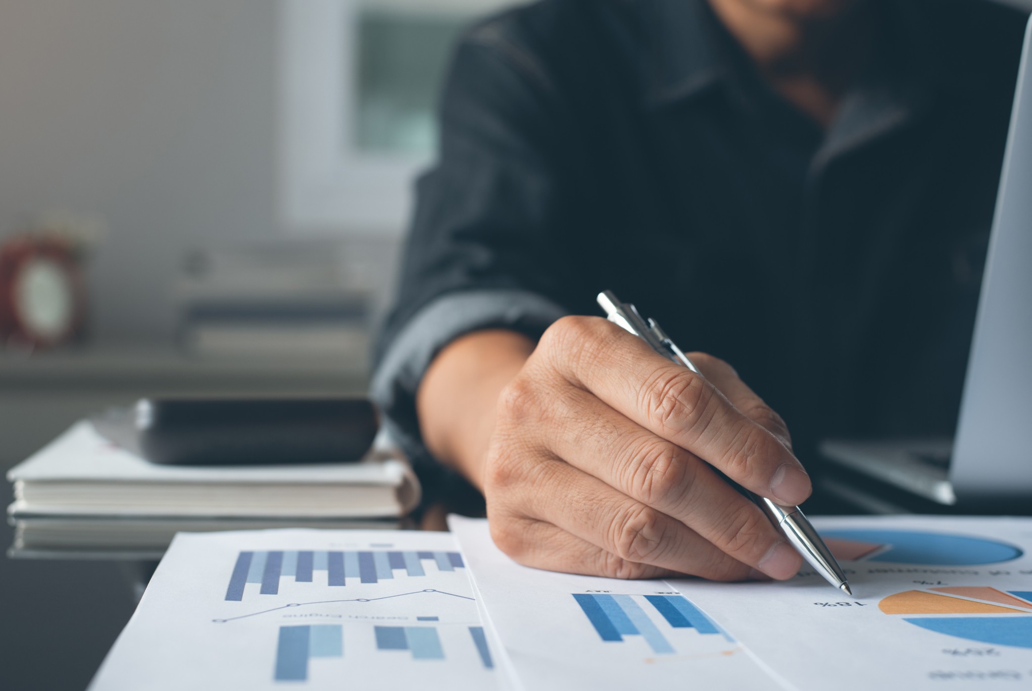 Close-up of a person analyzing financial charts and graphs with a pen, while working on a laptop, representing revenue cycle analytics in healthcare.