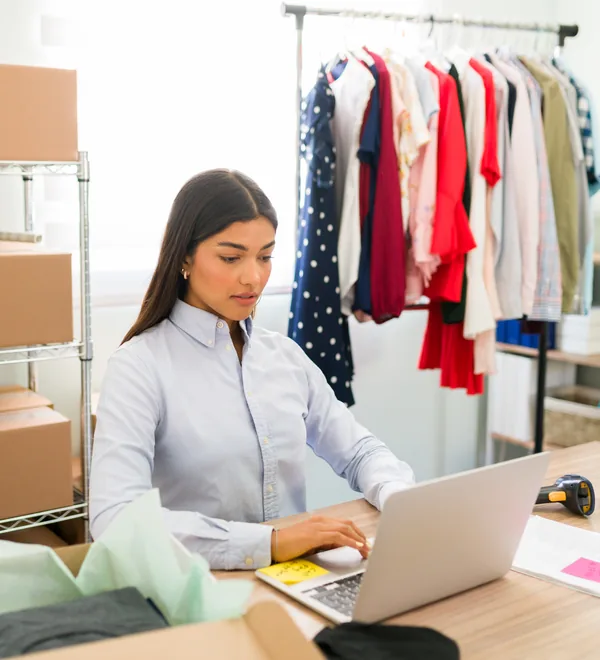 Apparel business owner answering buyer orders on a laptop beside packed boxes