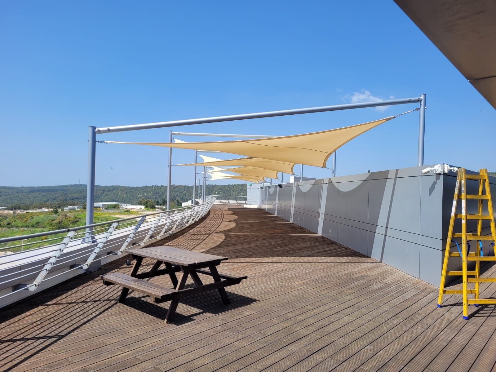 Wooden terrace with a picnic table; behind it, several triangular sun sails over a parapet against a clear sky.